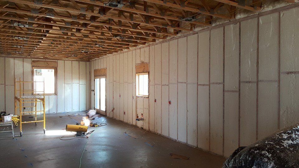Interior view of a building under construction, showing spray foam insulation on walls, with windows and unfinished ceiling.