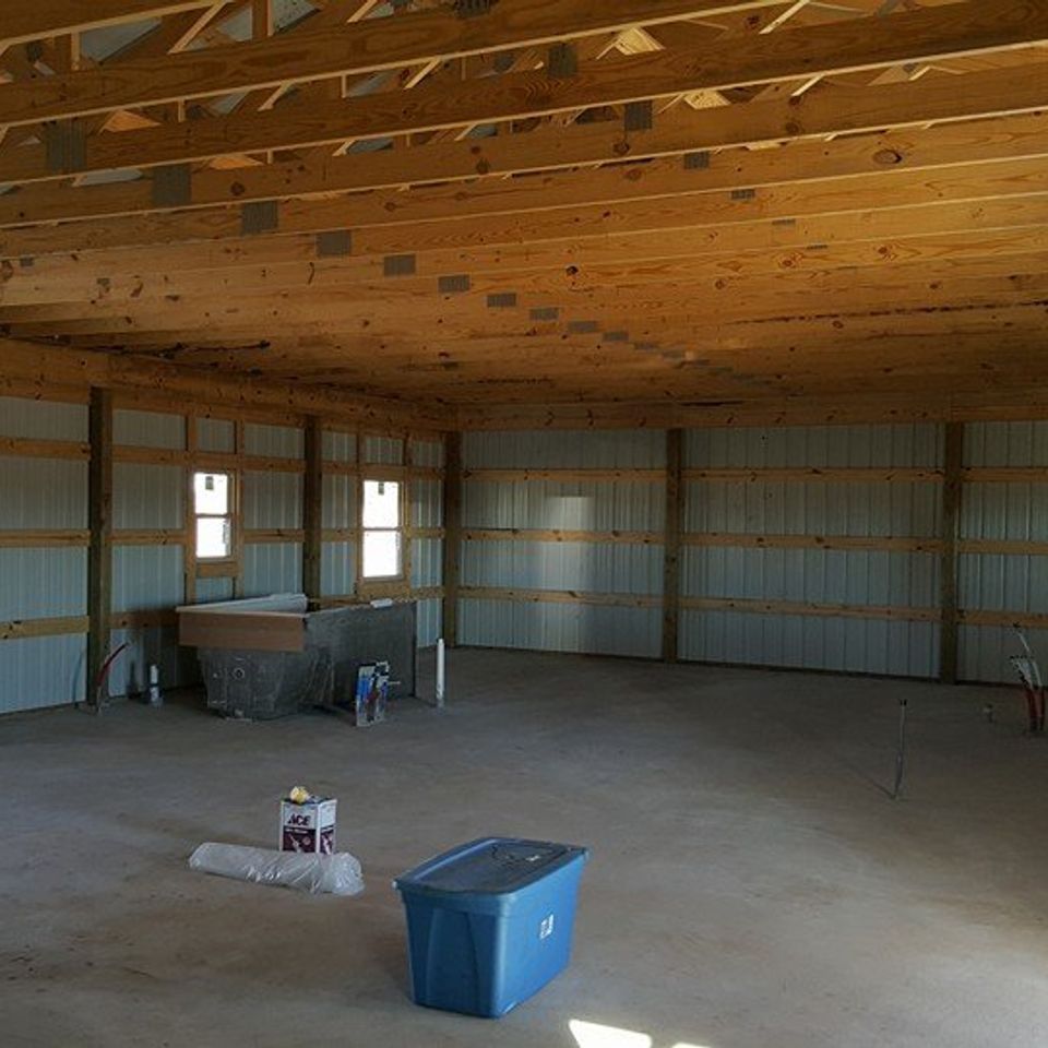 Interior of a large unfinished building with wooden beams, metal walls, and concrete floor; a blue bin sits in foreground.