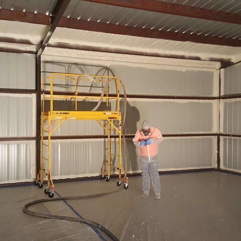 Person spraying insulation in a metal building, with scaffolding present.