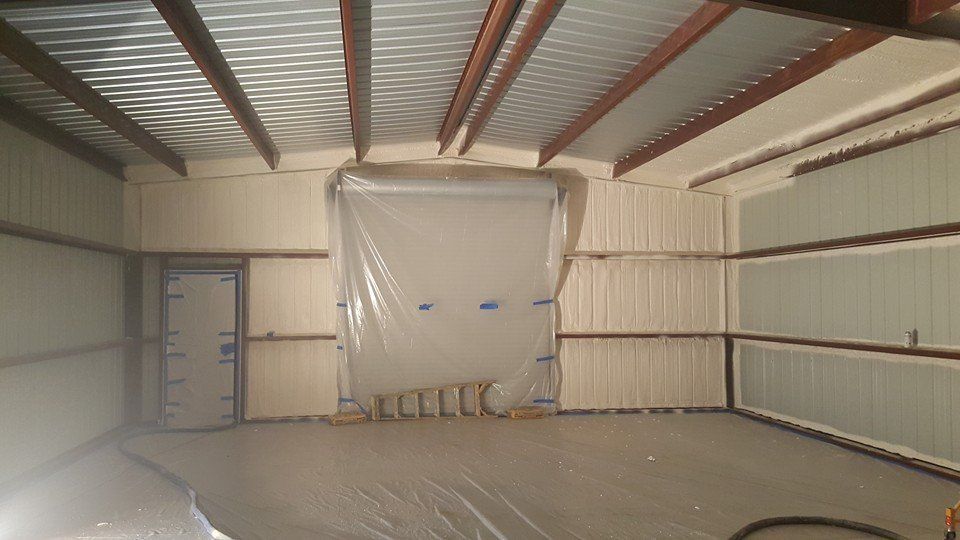 Interior of a metal building being insulated with spray foam. Walls, ceiling and door are covered in foam.