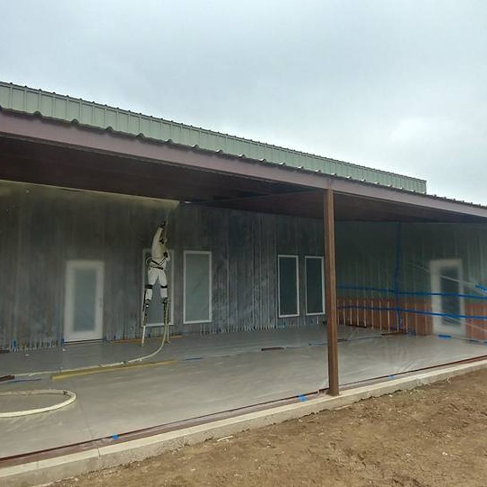A person in protective gear sprays paint on an exterior building, under a brown-roofed awning.