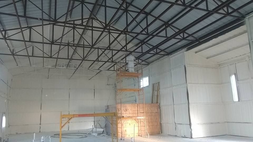 Interior of a building under construction, showing spray foam insulation on walls, metal roof truss, and scaffolding.