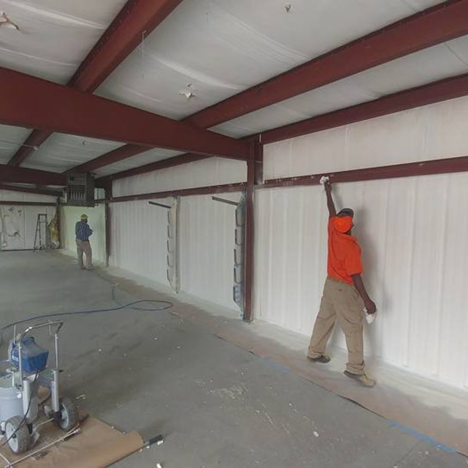 Workers spraying insulation on interior walls of a warehouse, wearing safety gear.