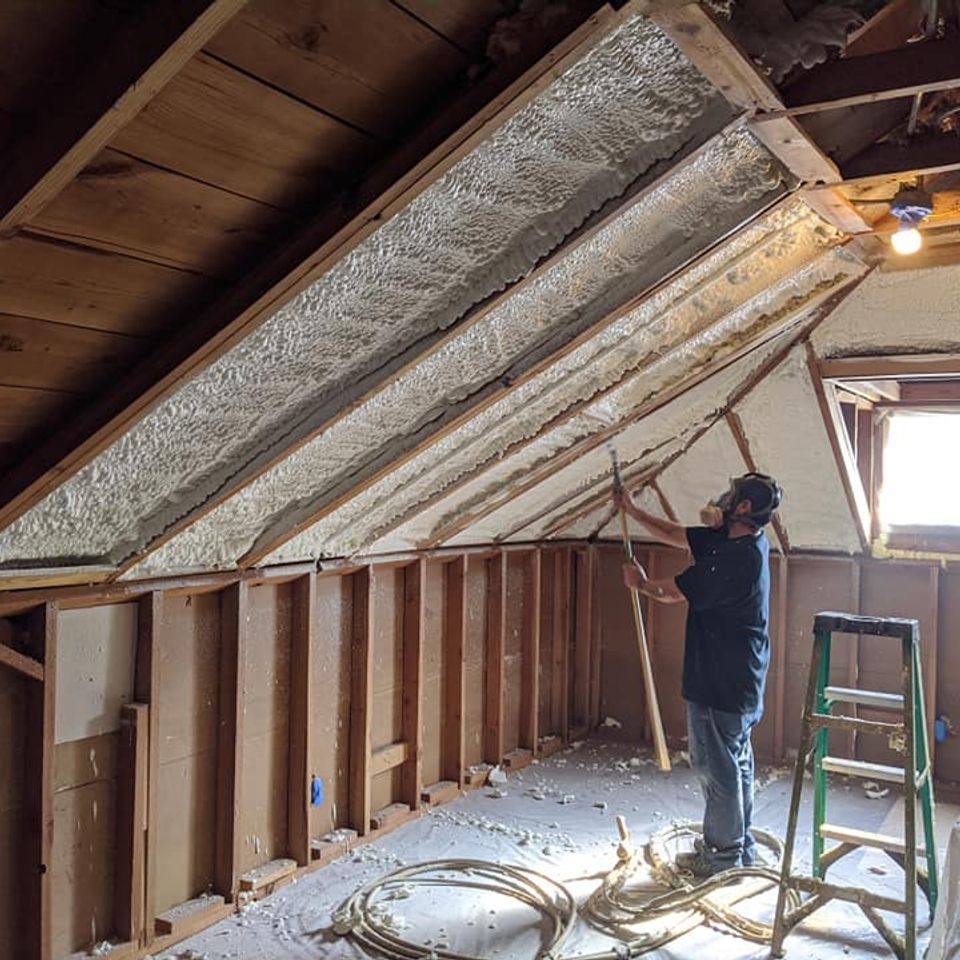 Man spraying foam insulation in an attic. Wooden beams, unfinished walls. Green ladder.