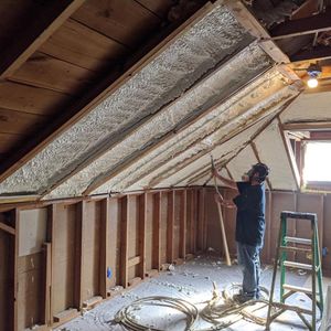 Man spraying foam insulation in an attic with exposed rafters. Walls are framed with wood.
