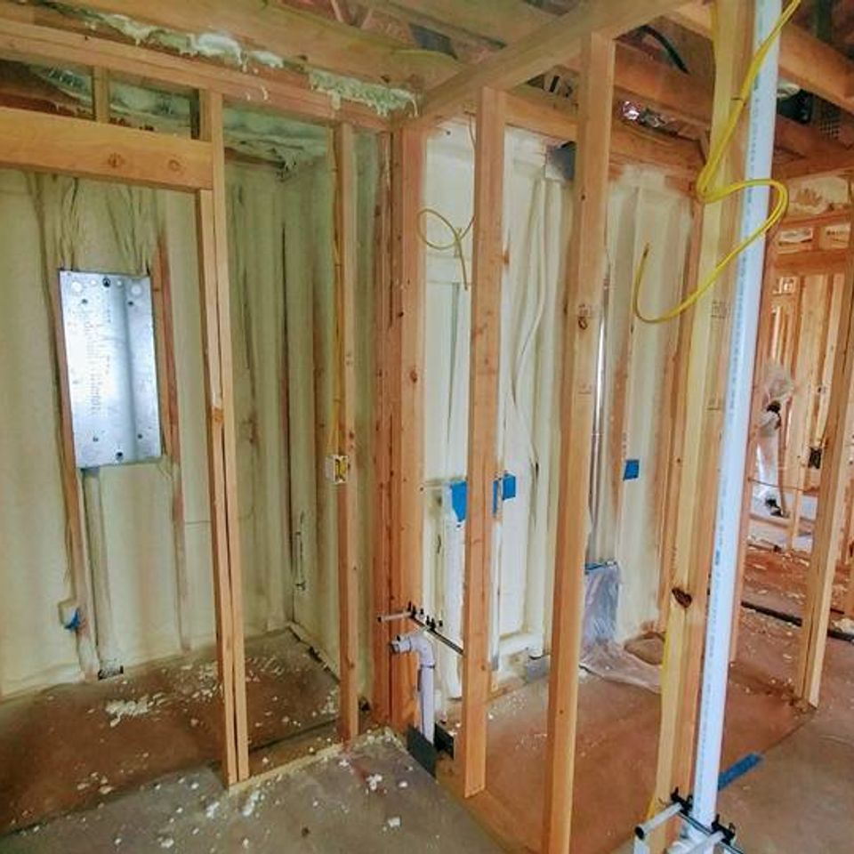 Interior view of a building's wooden frame walls with spray foam insulation applied. Wiring and plumbing visible.