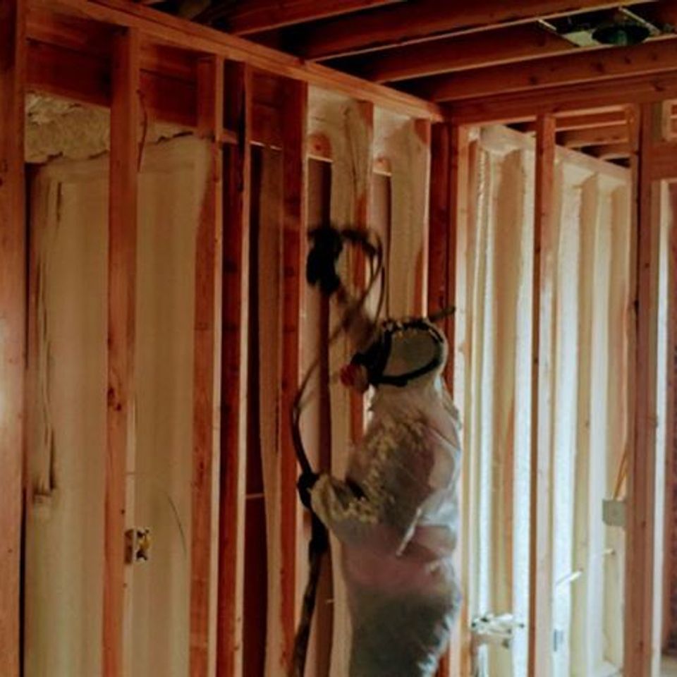Person in protective suit spraying foam insulation into wooden wall frame.