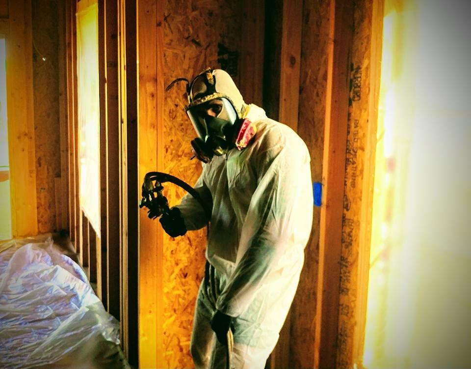 Person in protective suit and respirator sprays inside a wooden-framed structure.