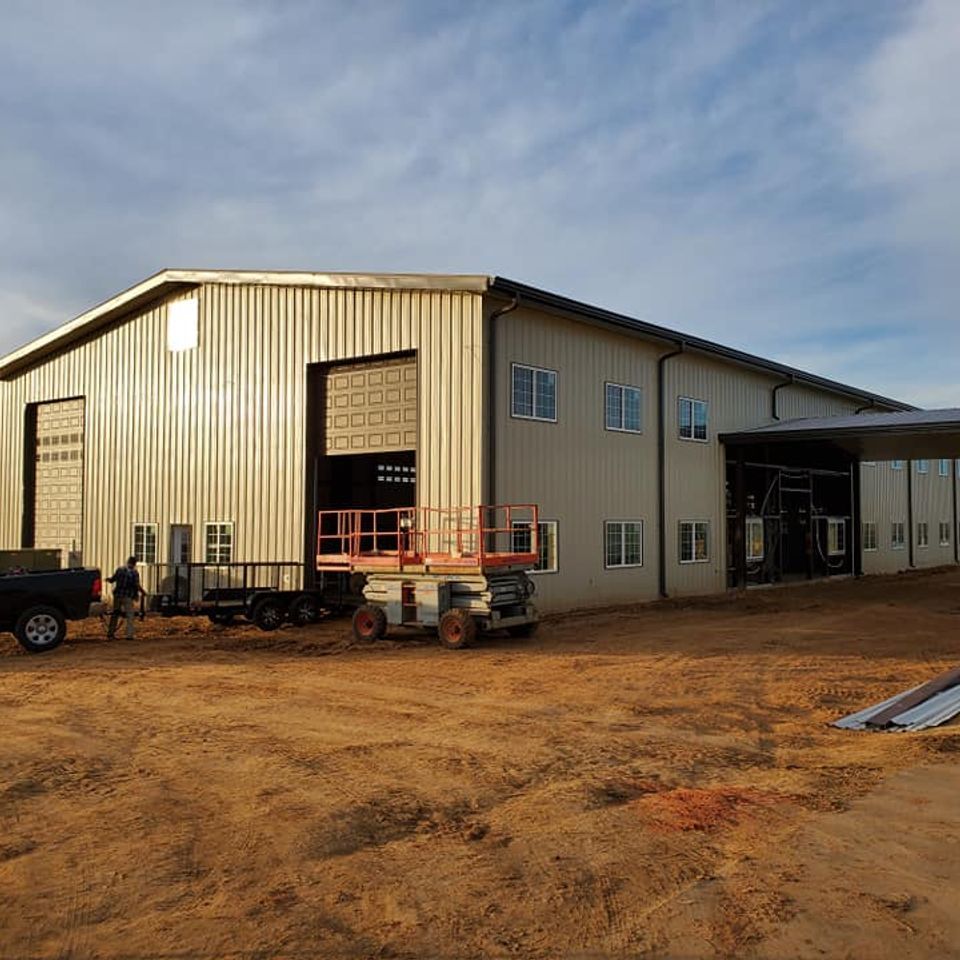 Large metal building under construction, with open bay door, lift, truck, trailer, and worker.
