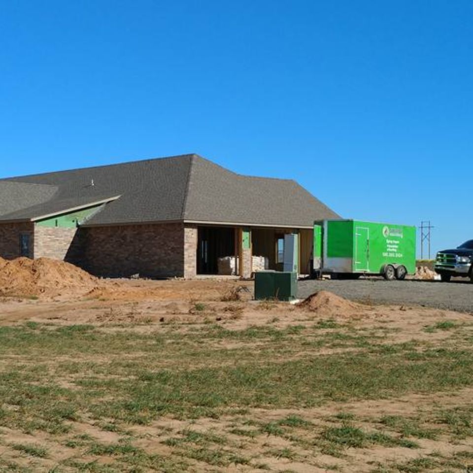 New home under construction; brick exterior, green trailer, dirt piles, clear blue sky.