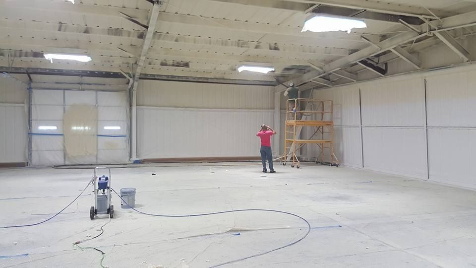 Man sprays paint in large, empty, white-walled warehouse. A paint machine is on the floor.