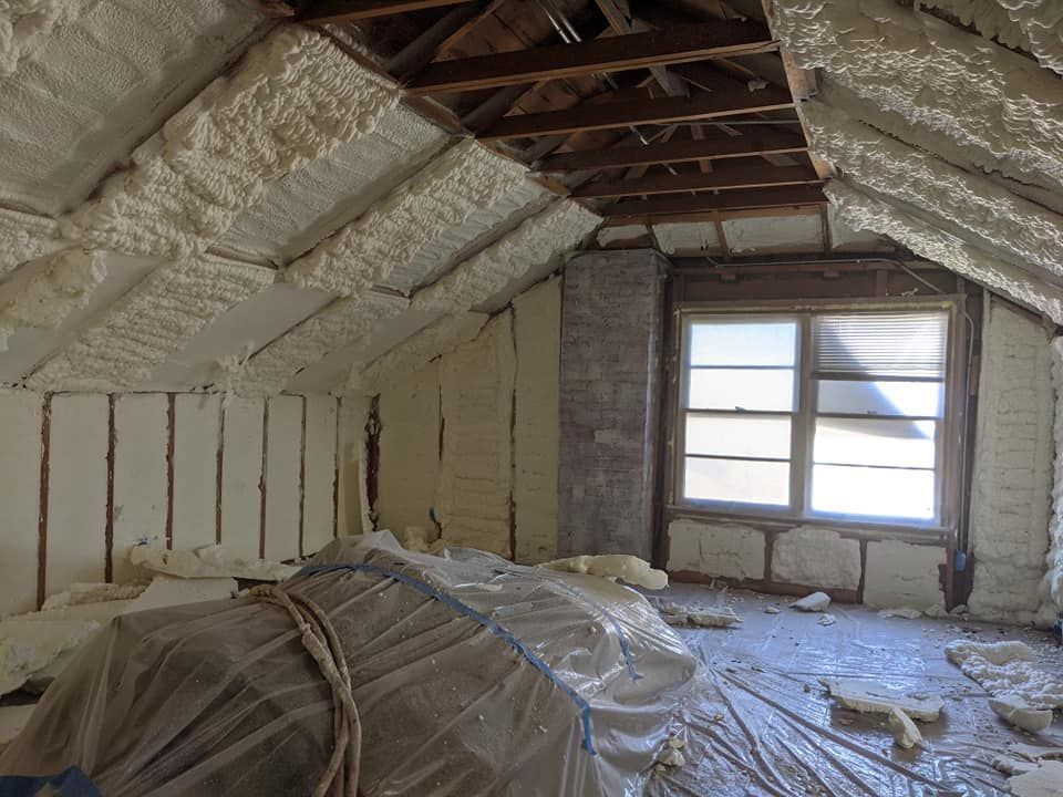 Attic room with spray foam insulation on walls and roof. Window in the back and tarp-covered items in the foreground.