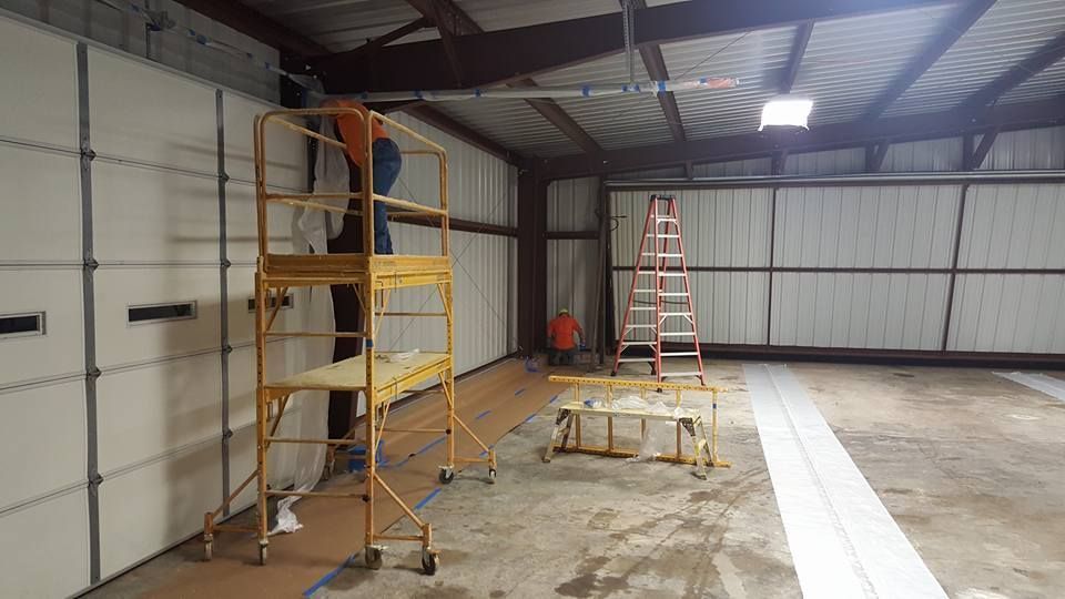 Person on a yellow scaffold working on a white wall inside a building, with a ladder and other materials visible.