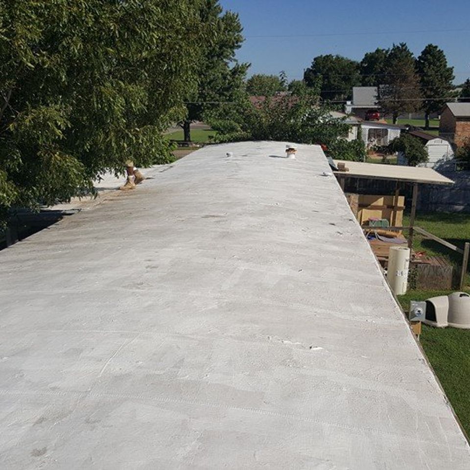 Flat, light gray roof of a building with a slightly sloped surface. Green trees and other buildings in the background.