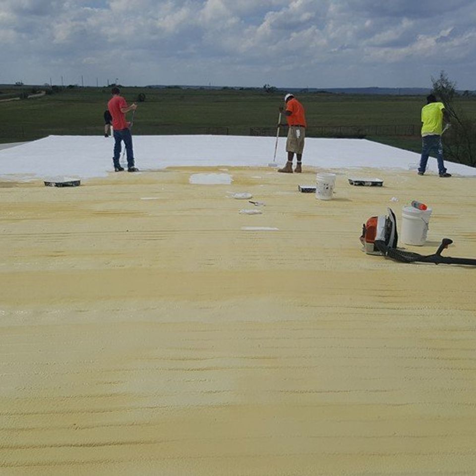 Three workers on a roof, applying a coating. Yellow roof, white coating area, green field background, cloudy sky.