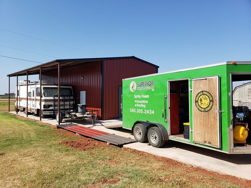 Green trailer and RV under a red metal building, trailer has company logo.