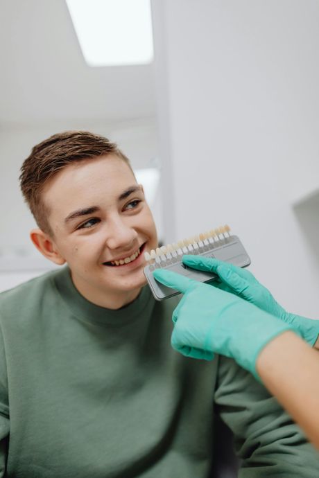 Person smiling while a gloved hand holds a dental shade guide near their teeth in a clinic
