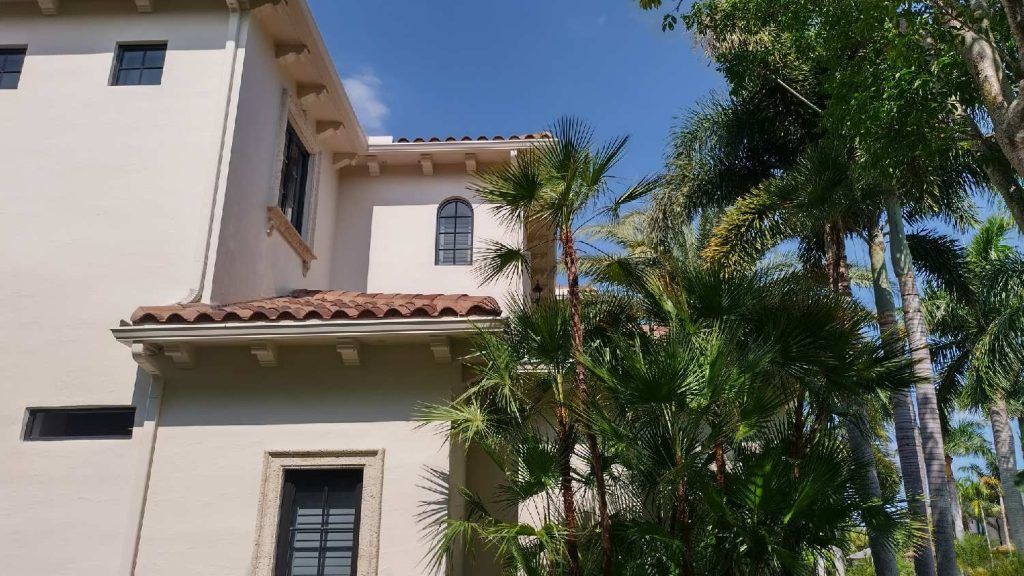 A large white house with a tiled roof is surrounded by palm trees on a sunny day.