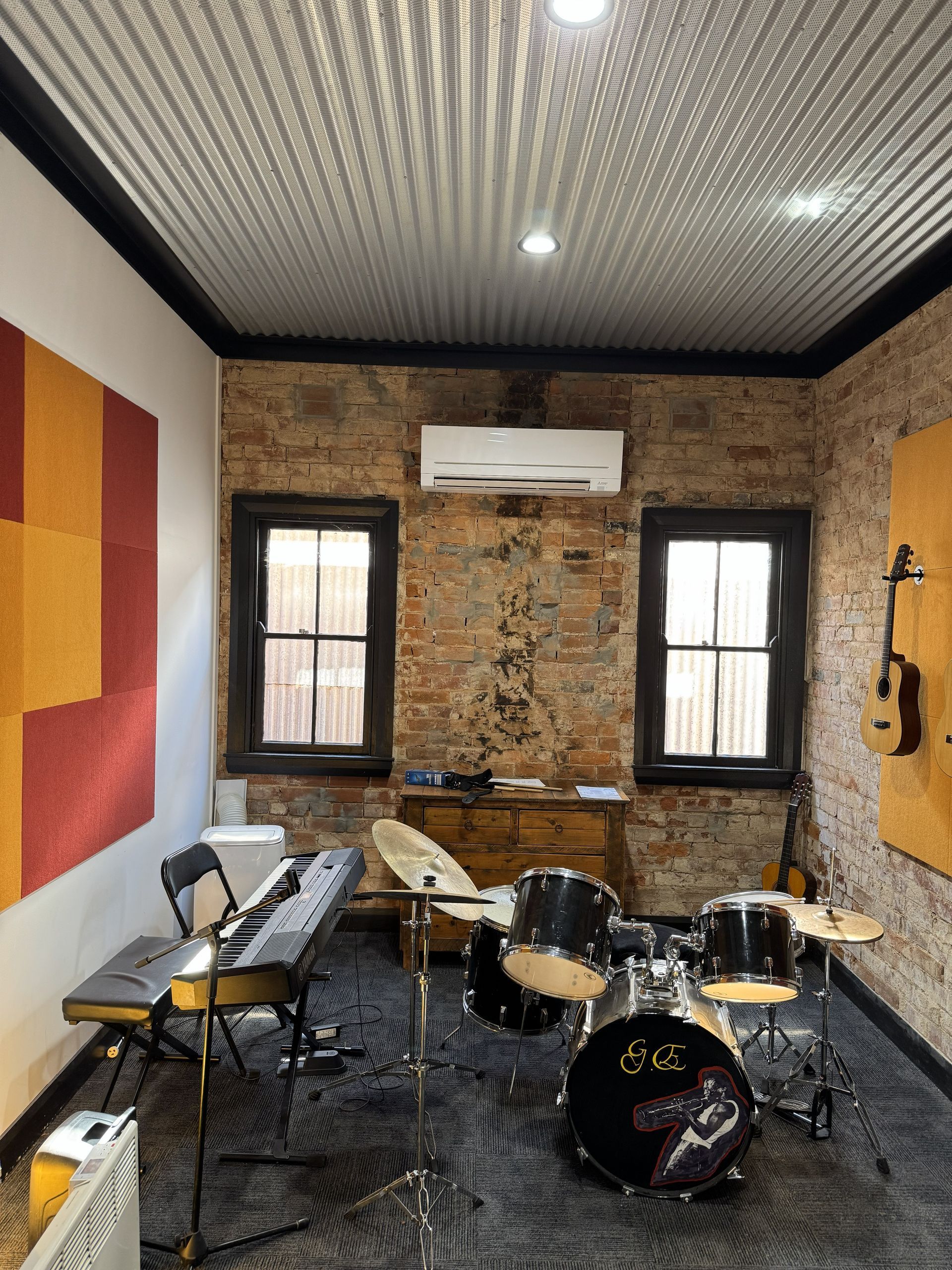 Music studio with drum set, keyboard, acoustic panels, brick walls, and corrugated ceiling.