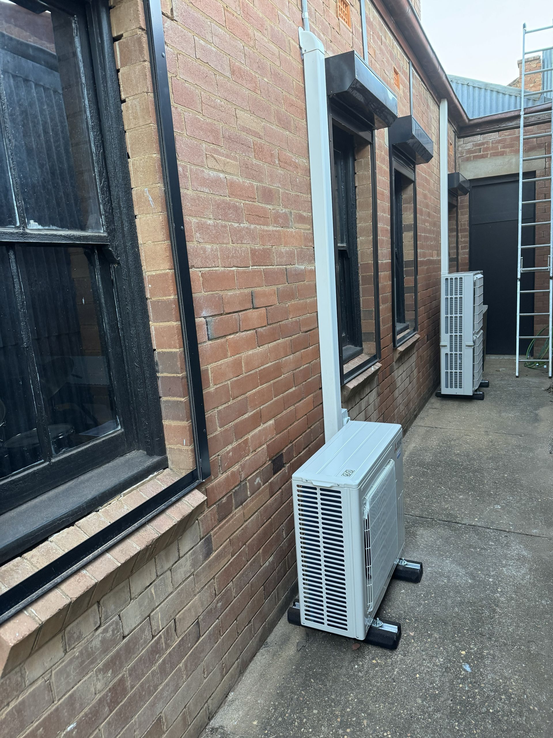 Air conditioning units sit outside against a brick building with black-framed windows.
