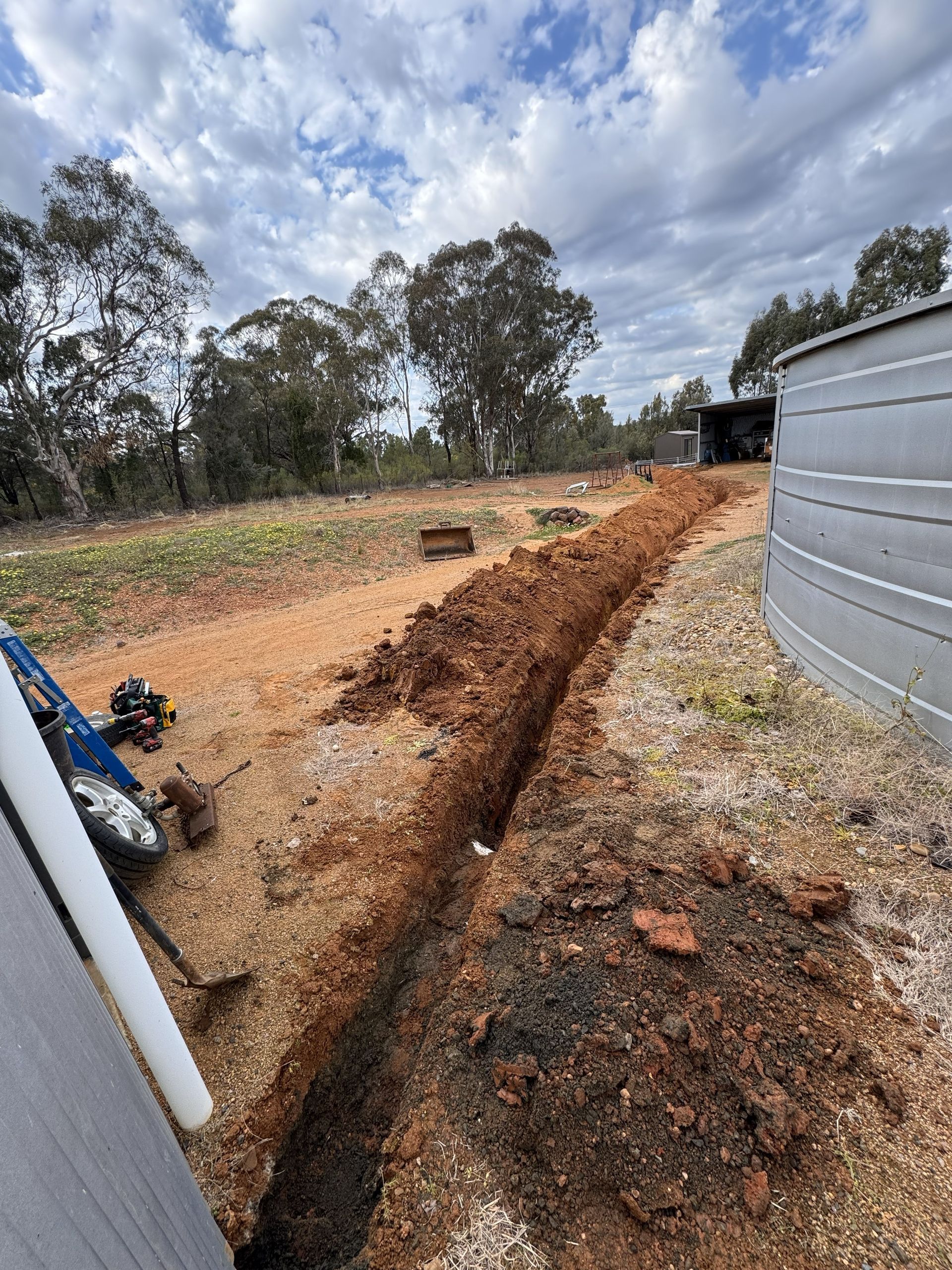 Trench dug in red dirt, next to a water tank. Overcast sky with trees in the background.