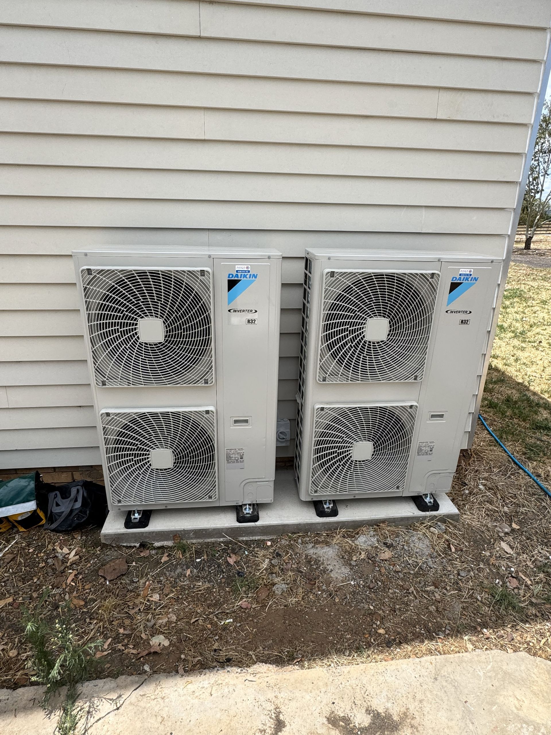 Two Daikin heat pump units installed on a concrete pad next to a house with beige siding.