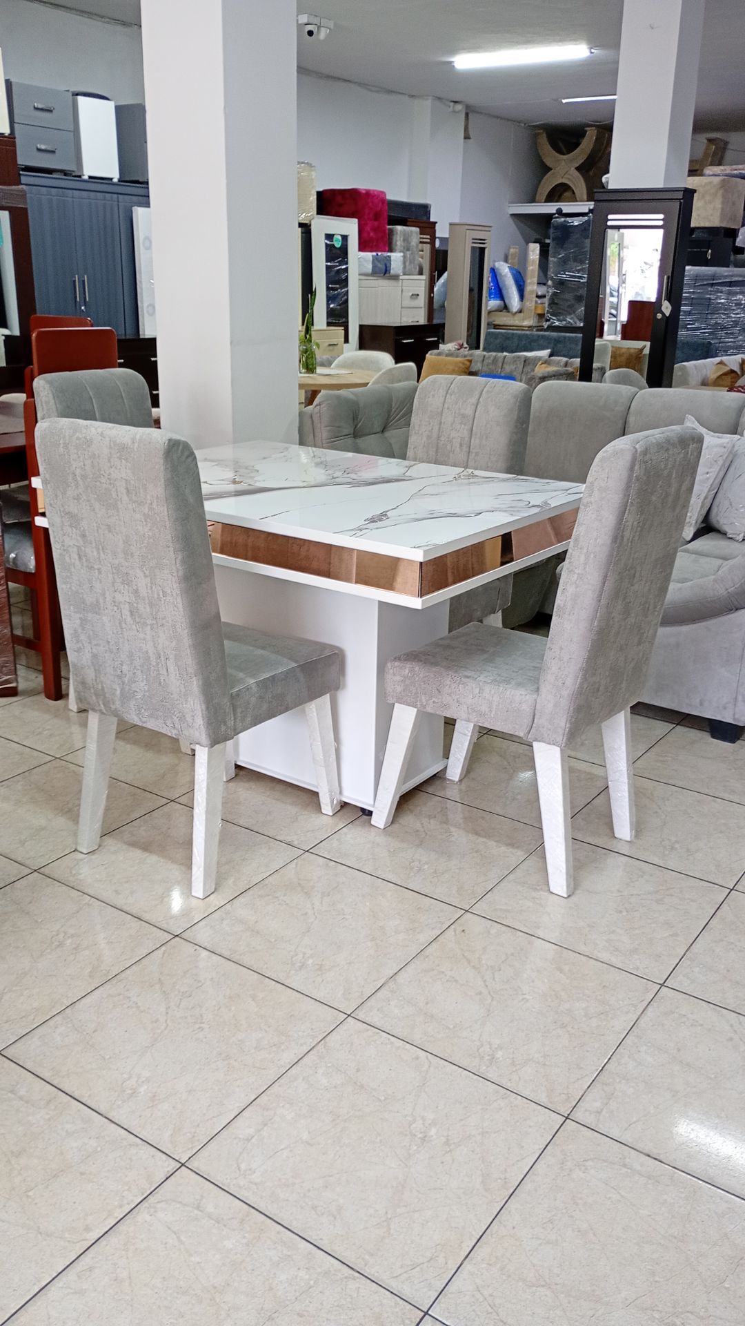 Mesa de comedor de exposición con sillas blancas sobre un suelo de baldosas, rodeada de muebles expuestos.