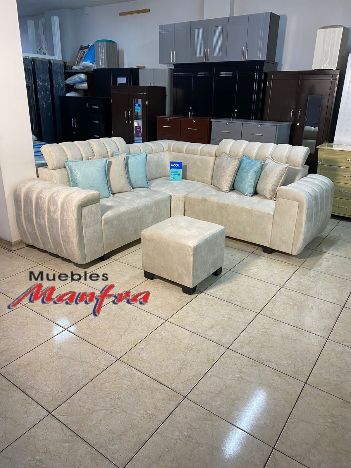 Sala de exposición de muebles modernos con un sofá seccional color crema, un puf a juego y armarios oscuros al fondo.