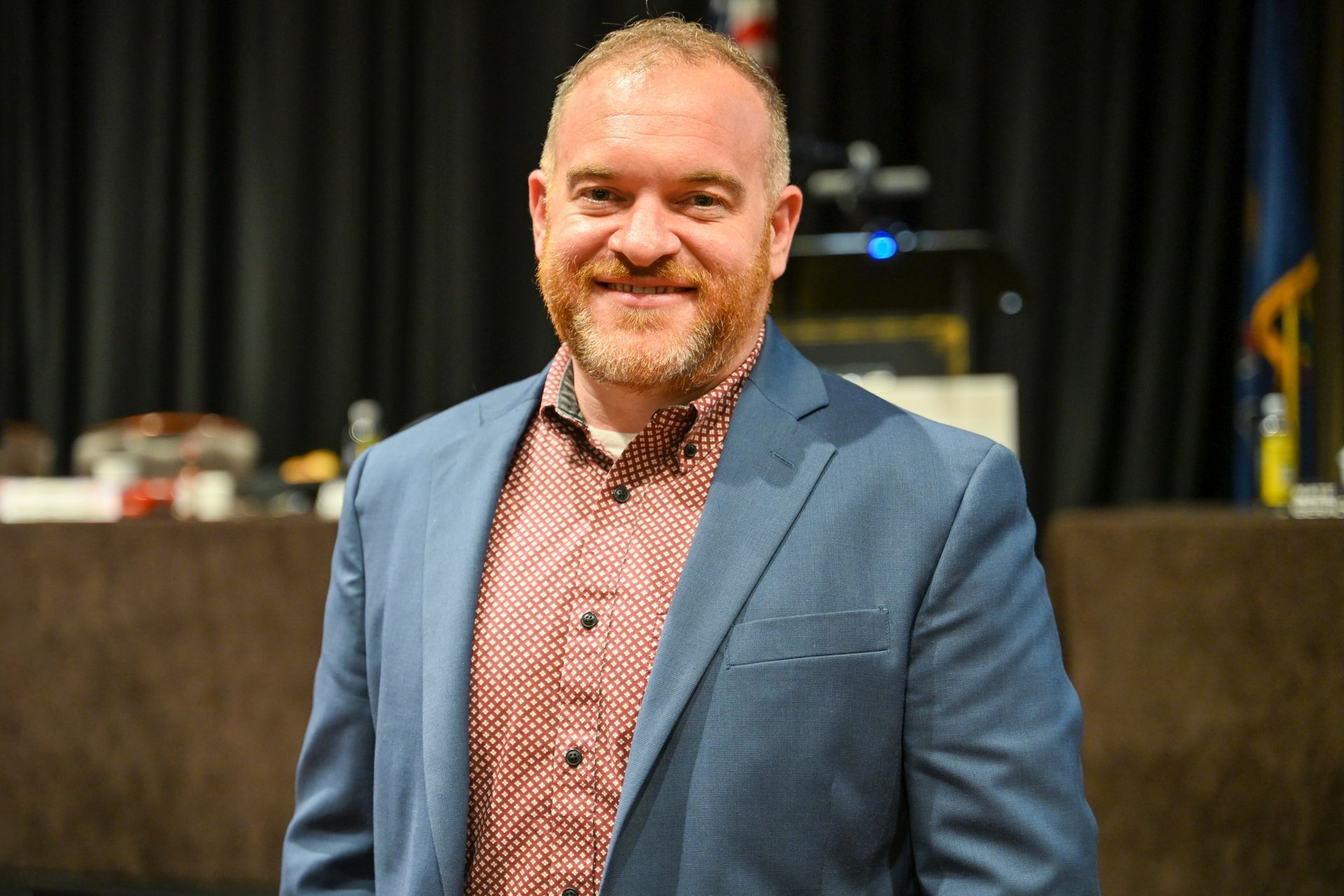 Man in blue blazer and red shirt smiling, standing in front of a dark backdrop.
