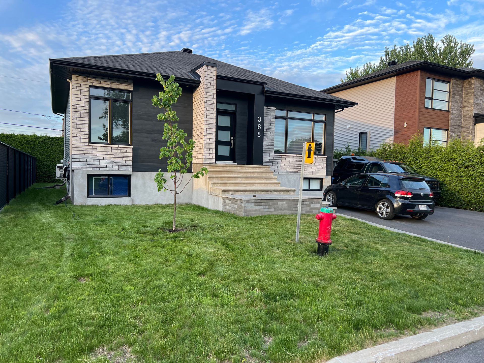 Maison moderne avec façade en pierre, marches d'entrée, cour gazonnée, voiture garée et borne d'incendie rouge.
