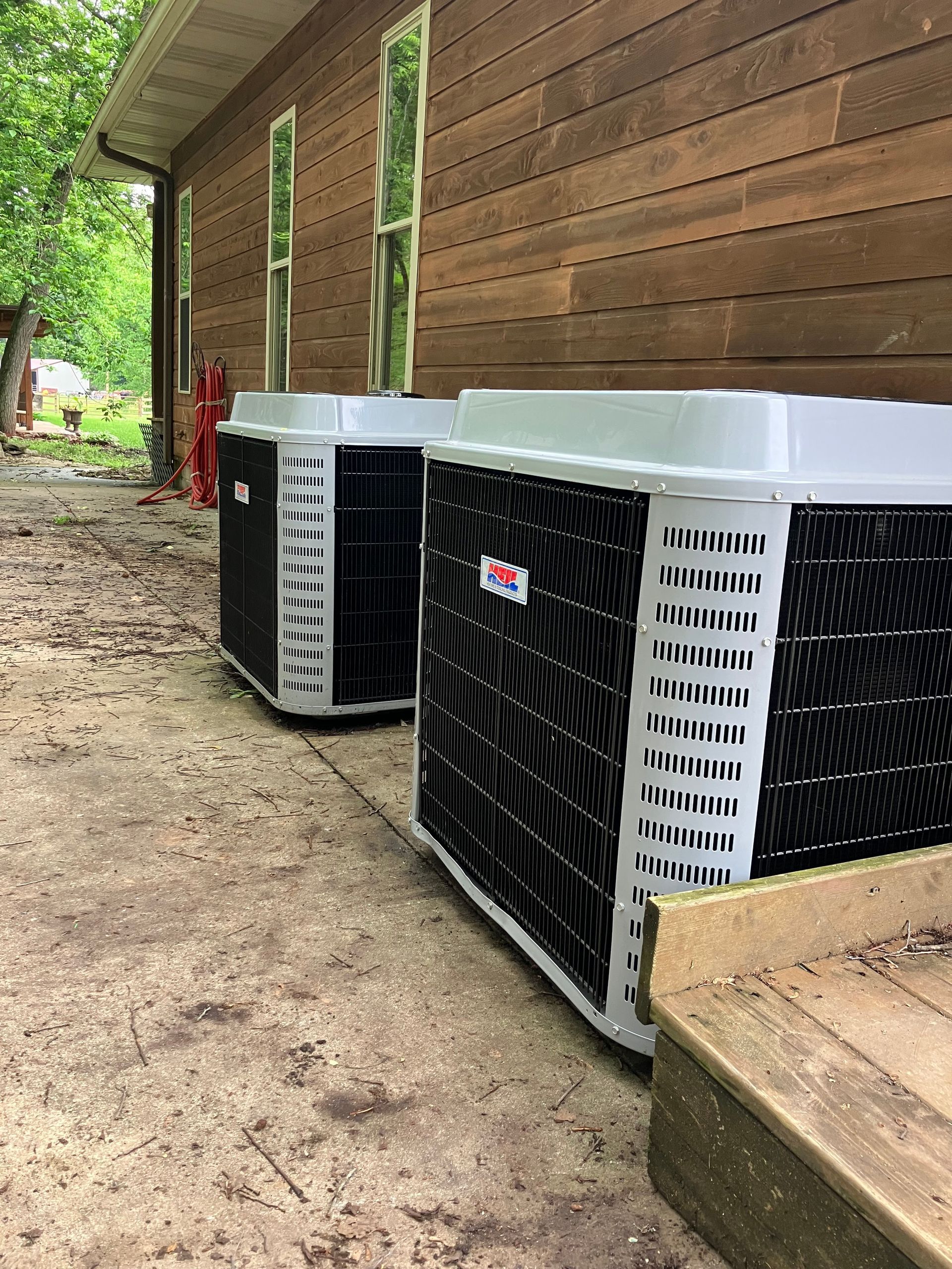 Two white outdoor HVAC units beside a wooden house on a dirt path