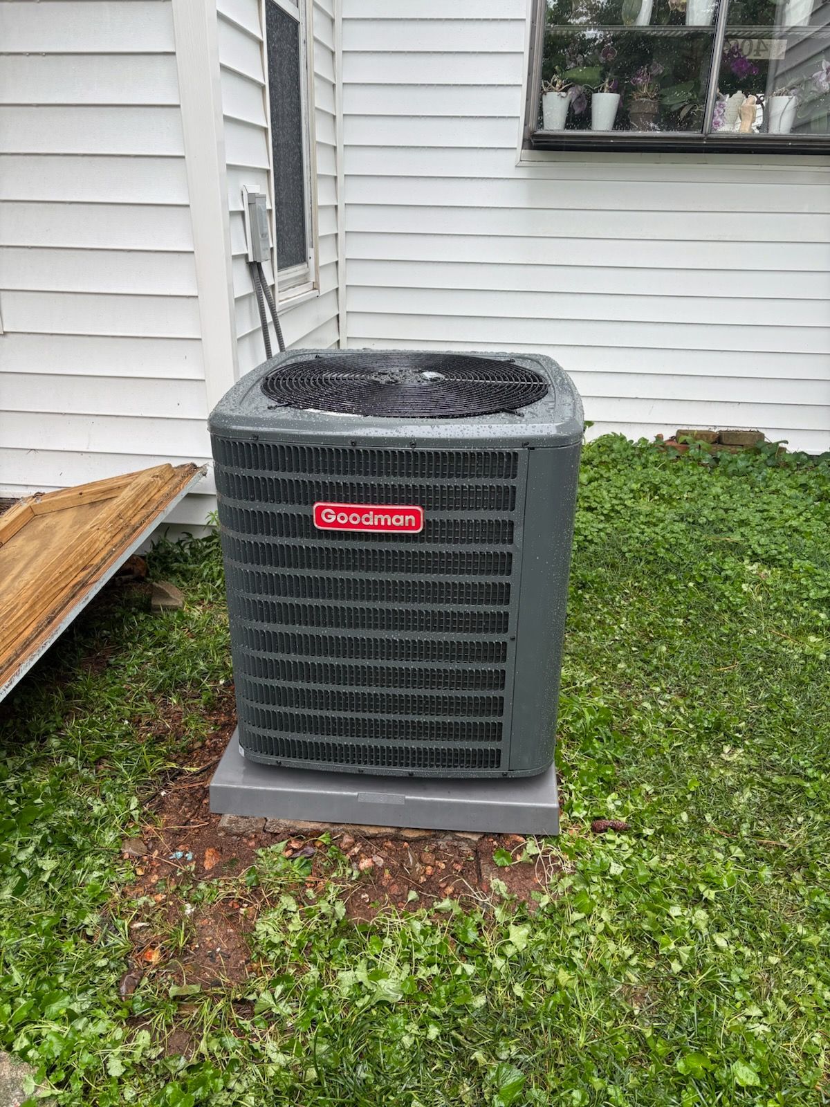 Outdoor air conditioner unit against a white house wall on a grassy lawn.