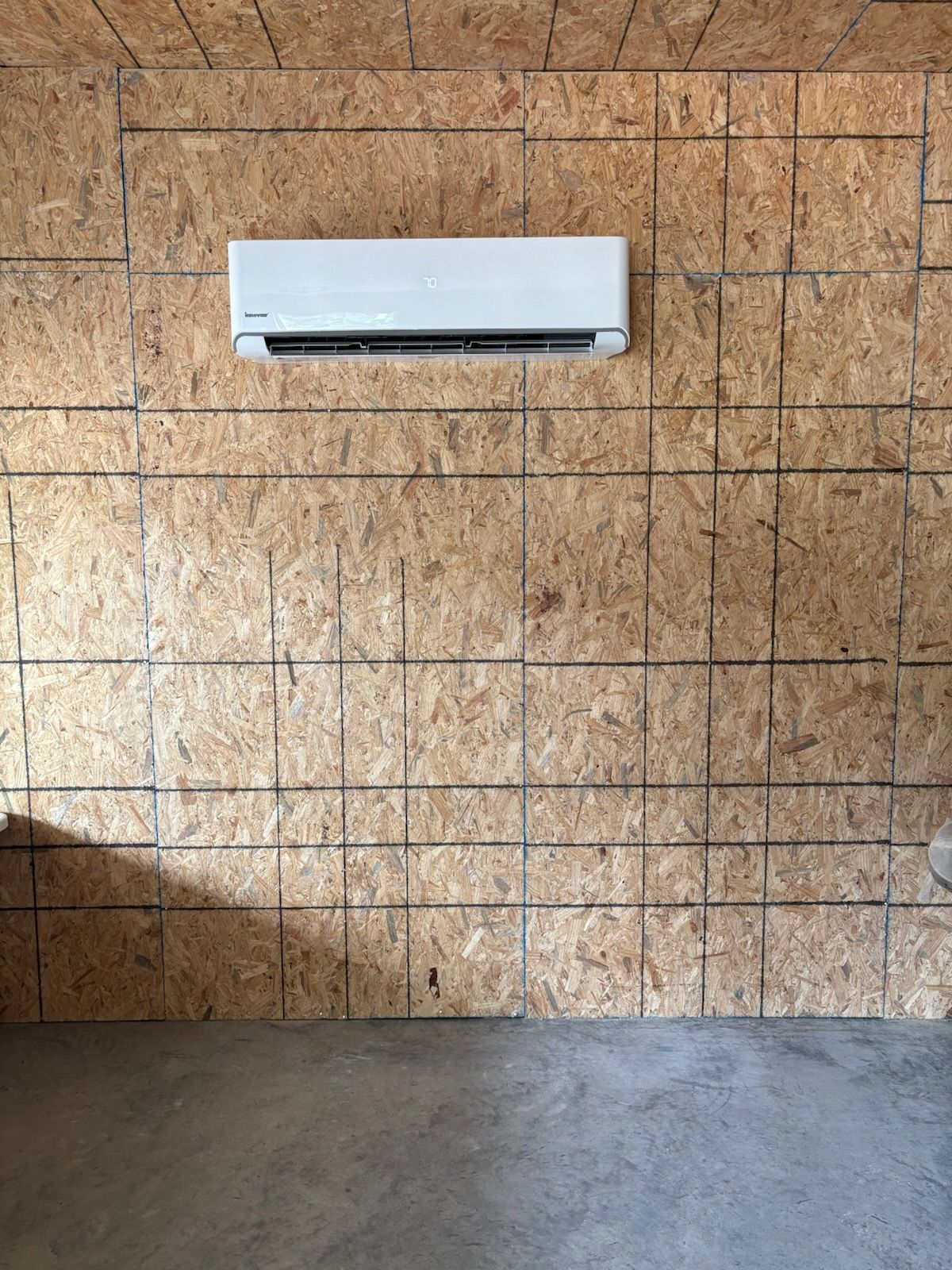 White wall-mounted air conditioner on a plywood wall above a concrete floor.