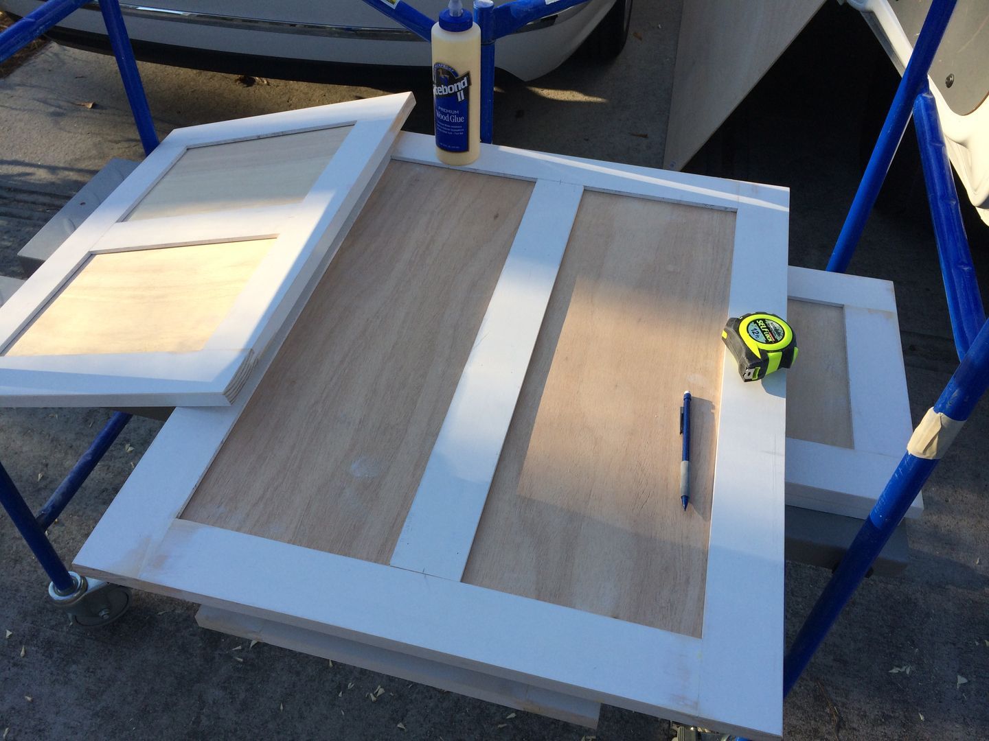 Wooden cabinet door panels on a blue cart, with wood glue and a tape measure.