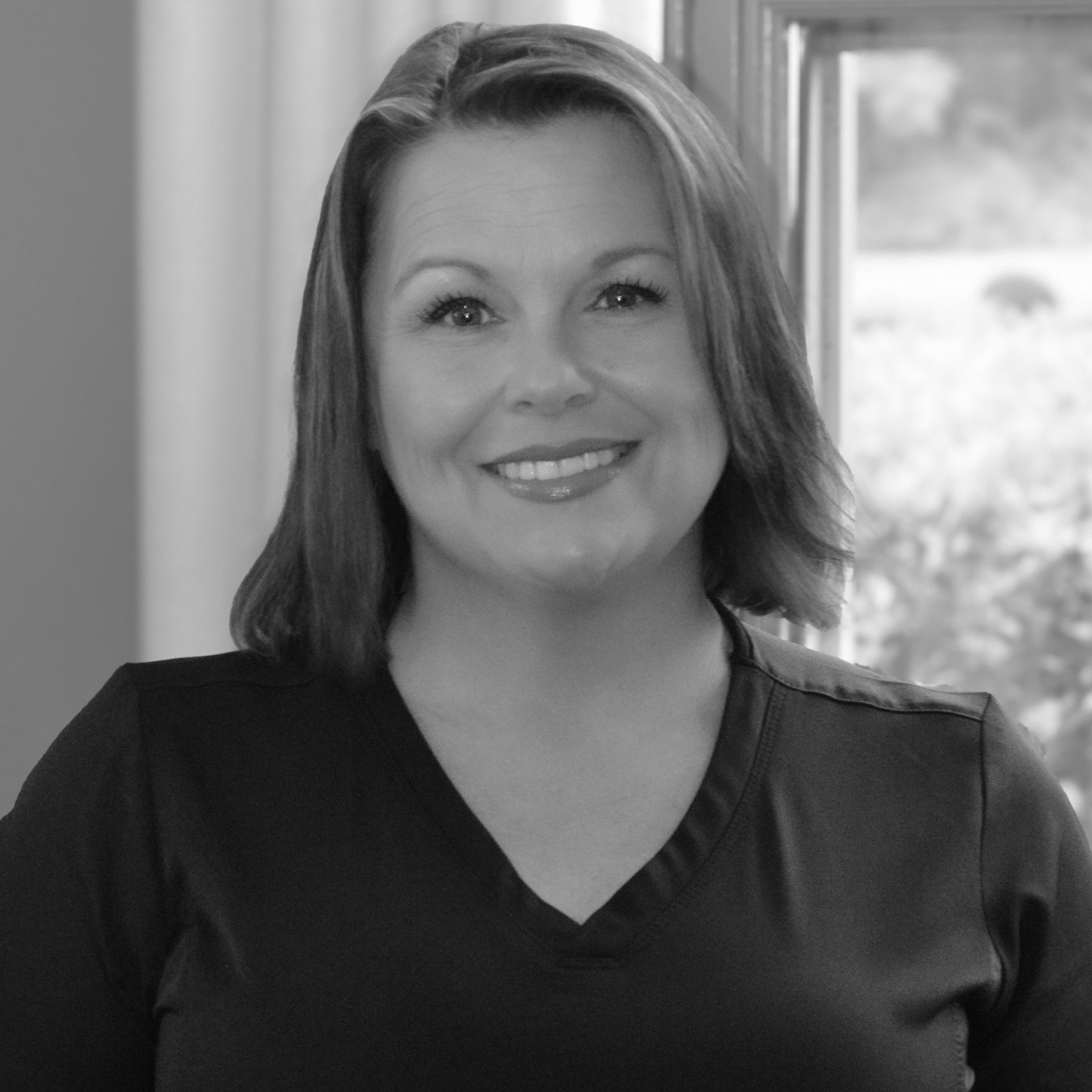 A woman in a black scrub top is smiling in front of a brick wall.