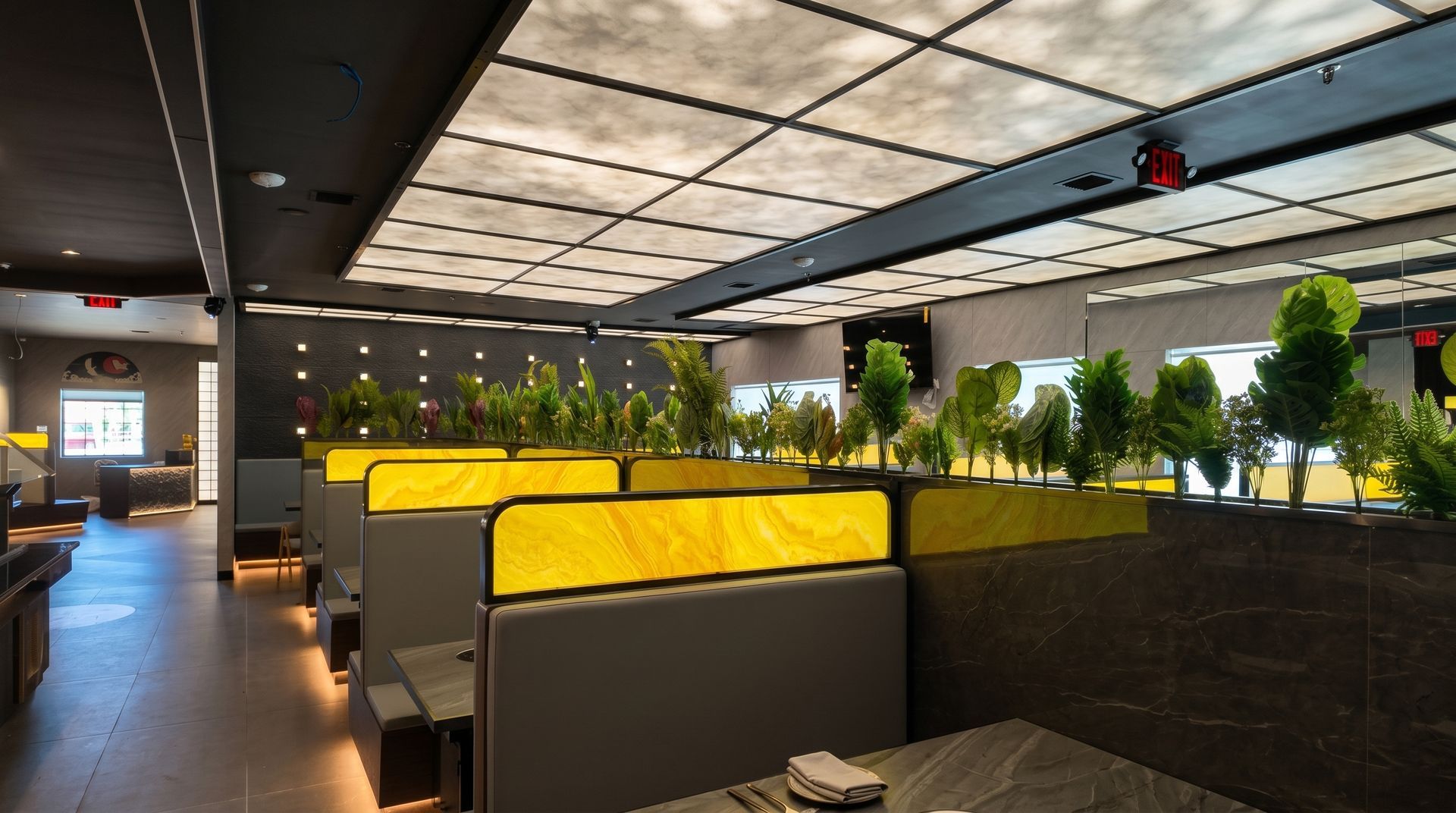 Modern office lounge with yellow booths, dark seating, and rows of potted plants under a lit ceiling