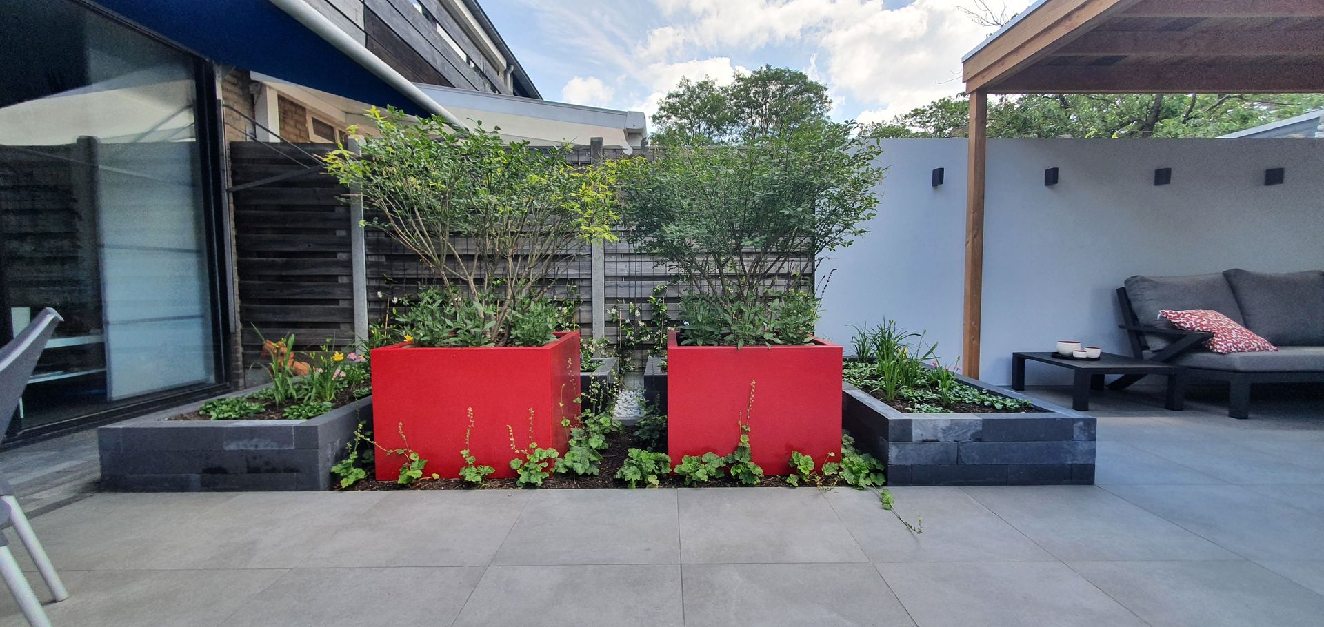 Modern terras met rode plantenbakken, grijze zitmeubelen en een zwarte bank tegen een witte muur.