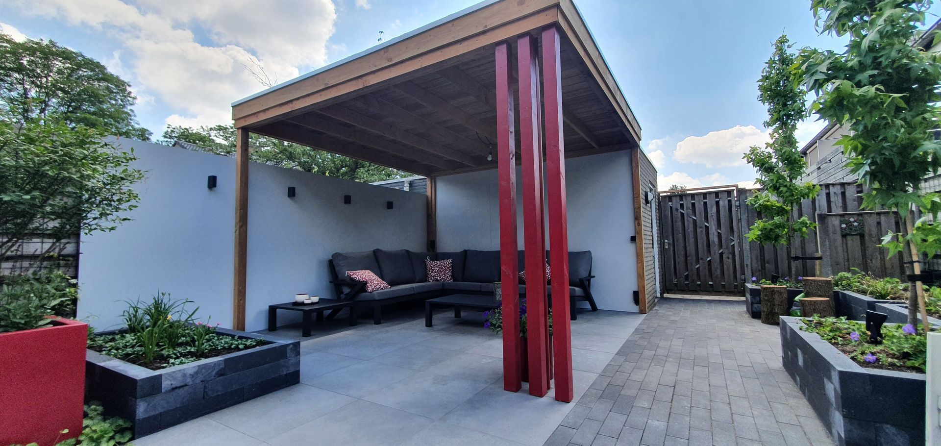 Moderne achtertuin met pergola, rode palen, tuinmeubilair, potplanten en bestrating.