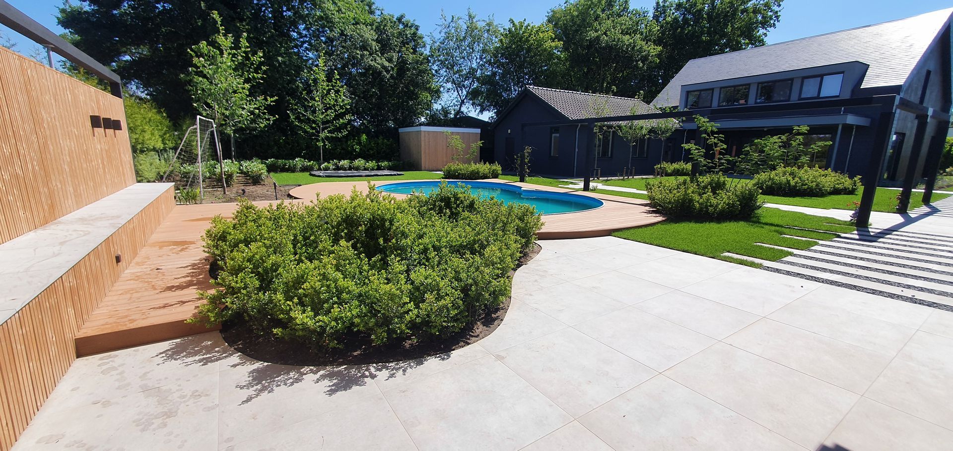 Achtertuin met zwembad, terras, groene struiken en een modern zwart huis in het volle zonlicht.