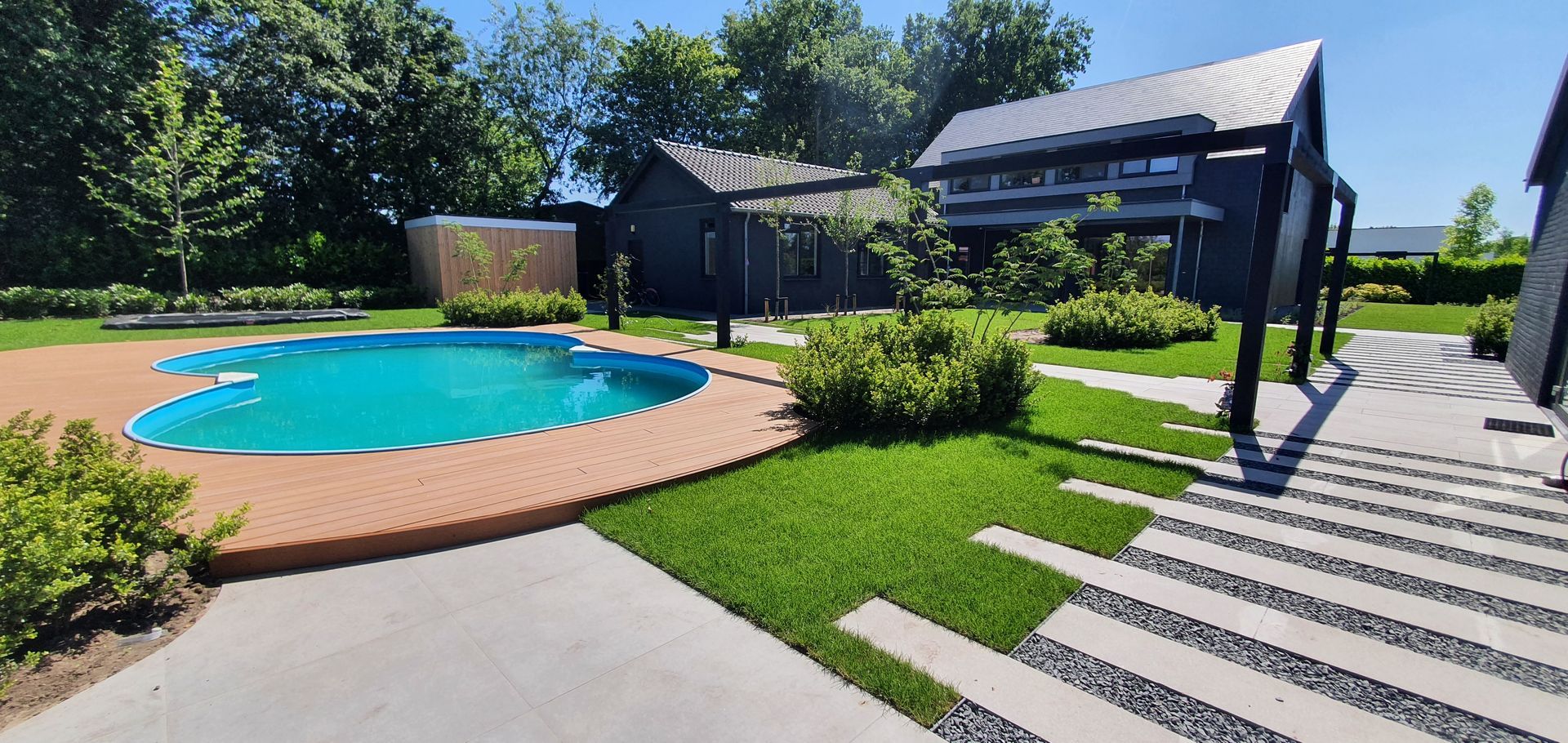 Buitenbad naast een aangelegde patio en een gestreept wandelpad bij een modern gebouw onder een blauwe hemel.