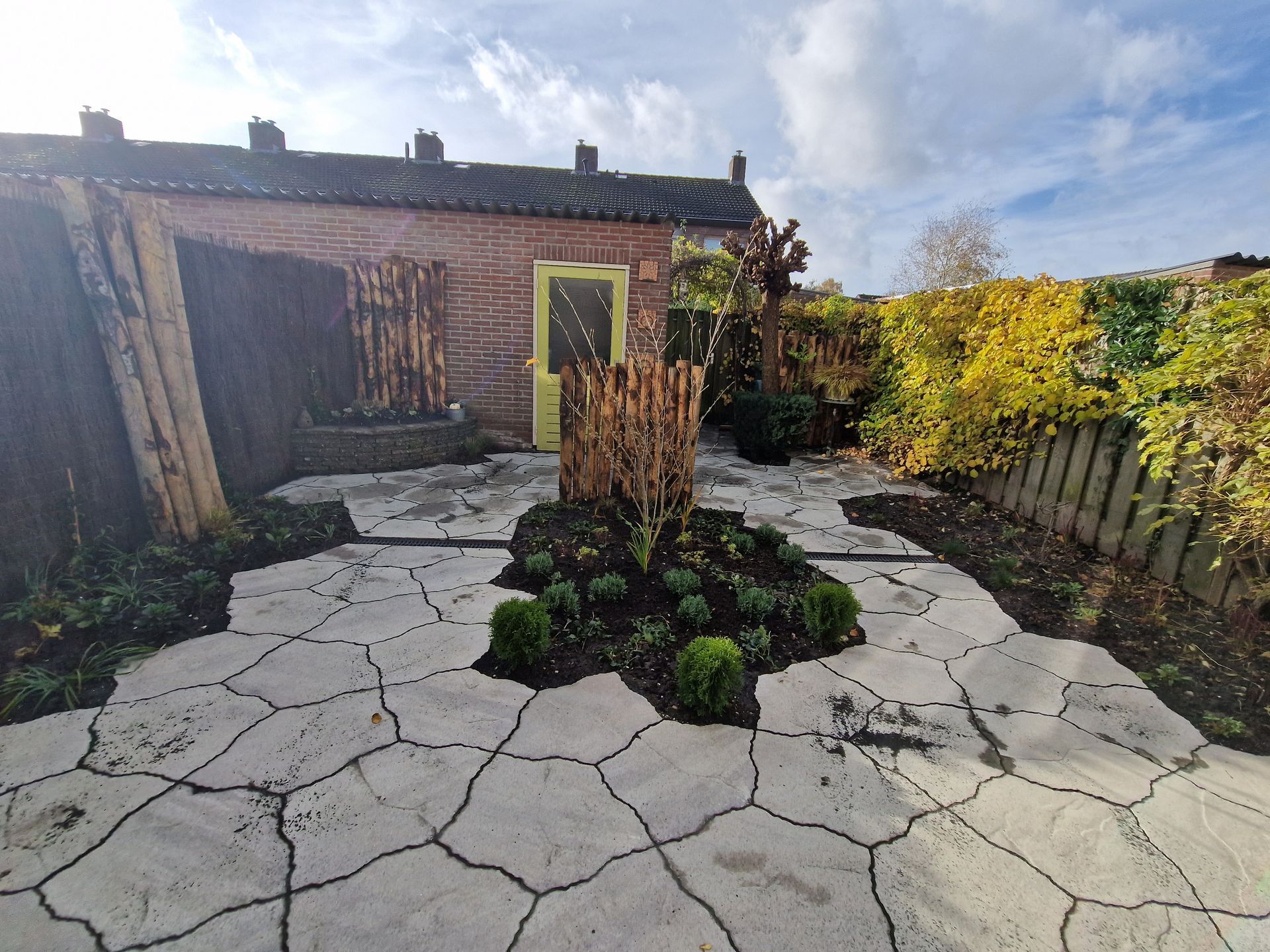 Een terras van gebroken beton met een centraal plantenbed en een gele deur in een rustiek bakstenen gebouw.
