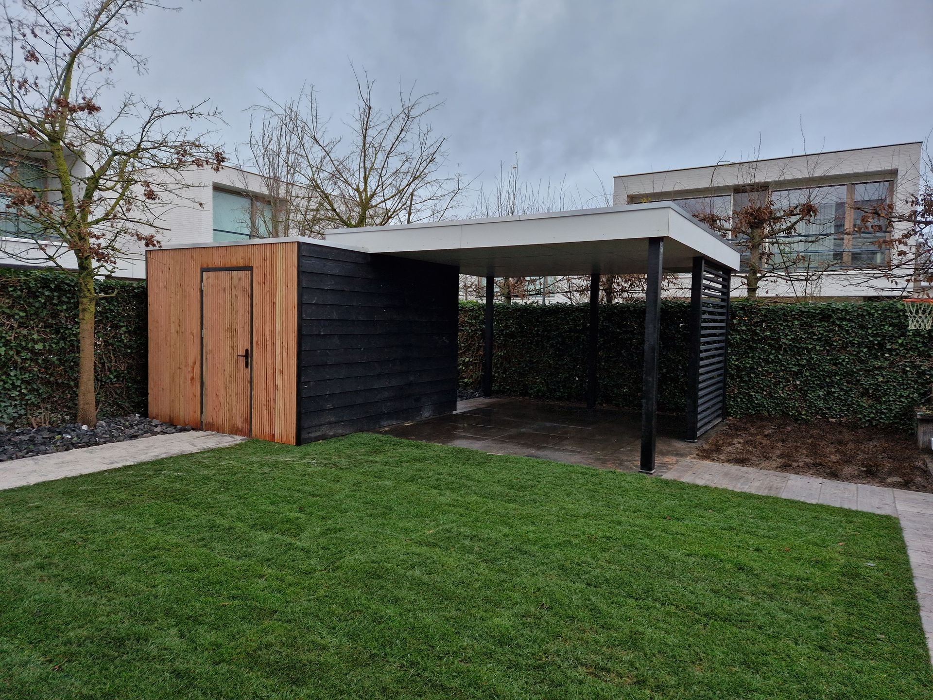 Een kleine achtertuin met een zwart-houten schuurtje, een overdekt terras en groen gras onder een bewolkte hemel.