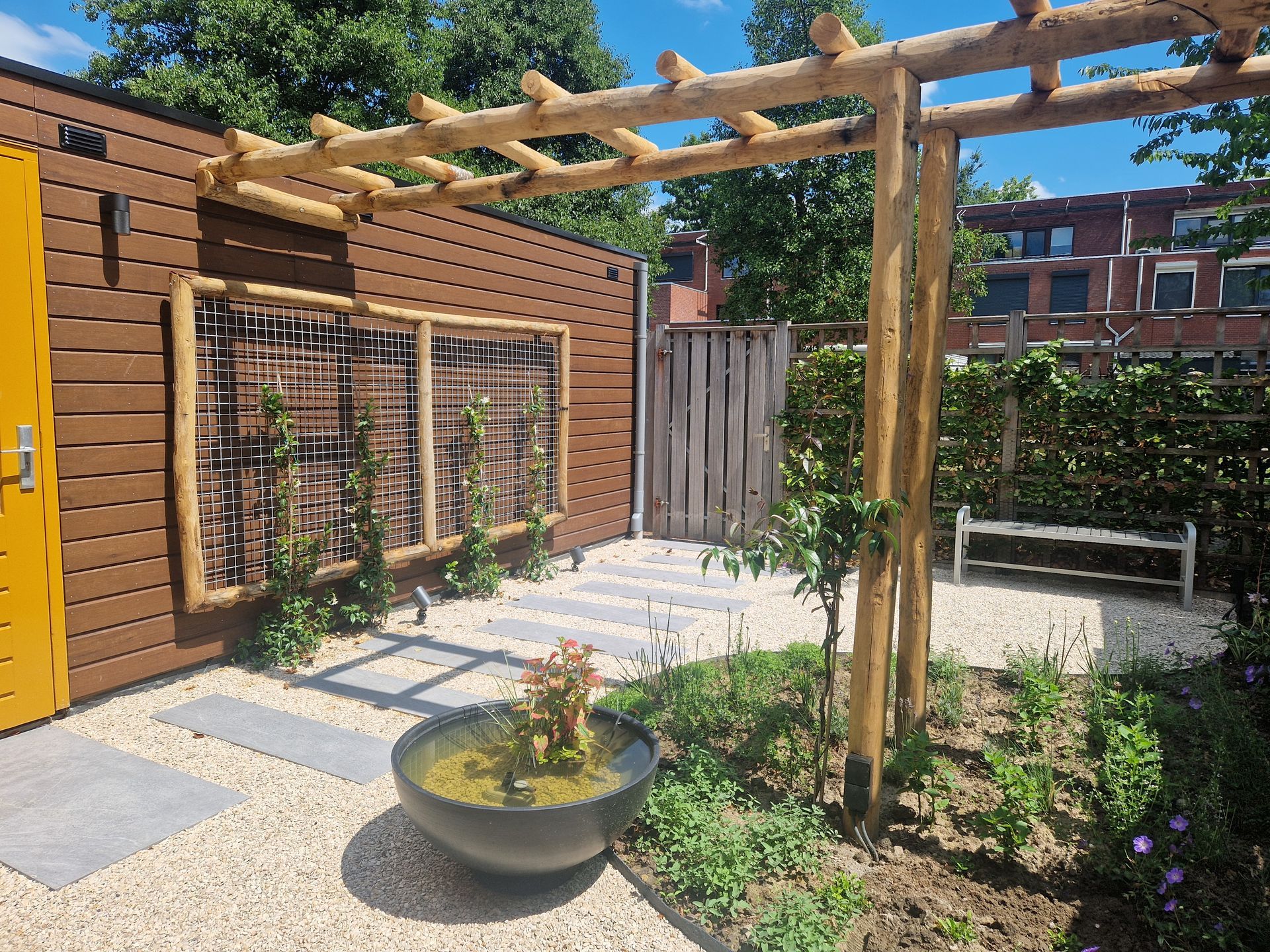 Zonnige tuin met houten pergola, bakstenen muur, stapstenen en een ronde plantenbak.