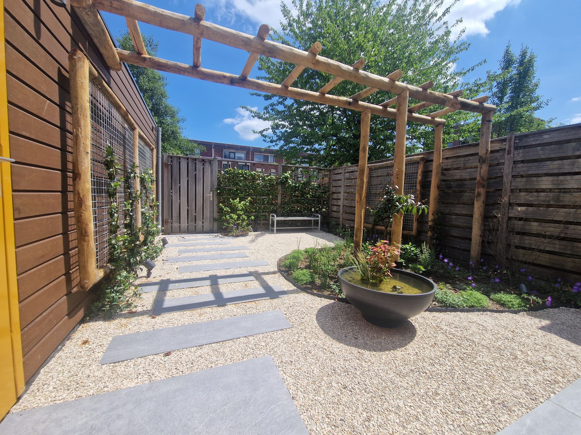 Terras in de achtertuin met grind, houten pergola, verhoogde plantenbak en omheinde zithoek in de zon.