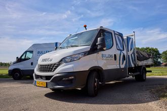 Een persoon in zwarte werkkleding knielt in een tuinbed van aarde en werkt vlakbij een bakstenen muur en zwaar materieel.