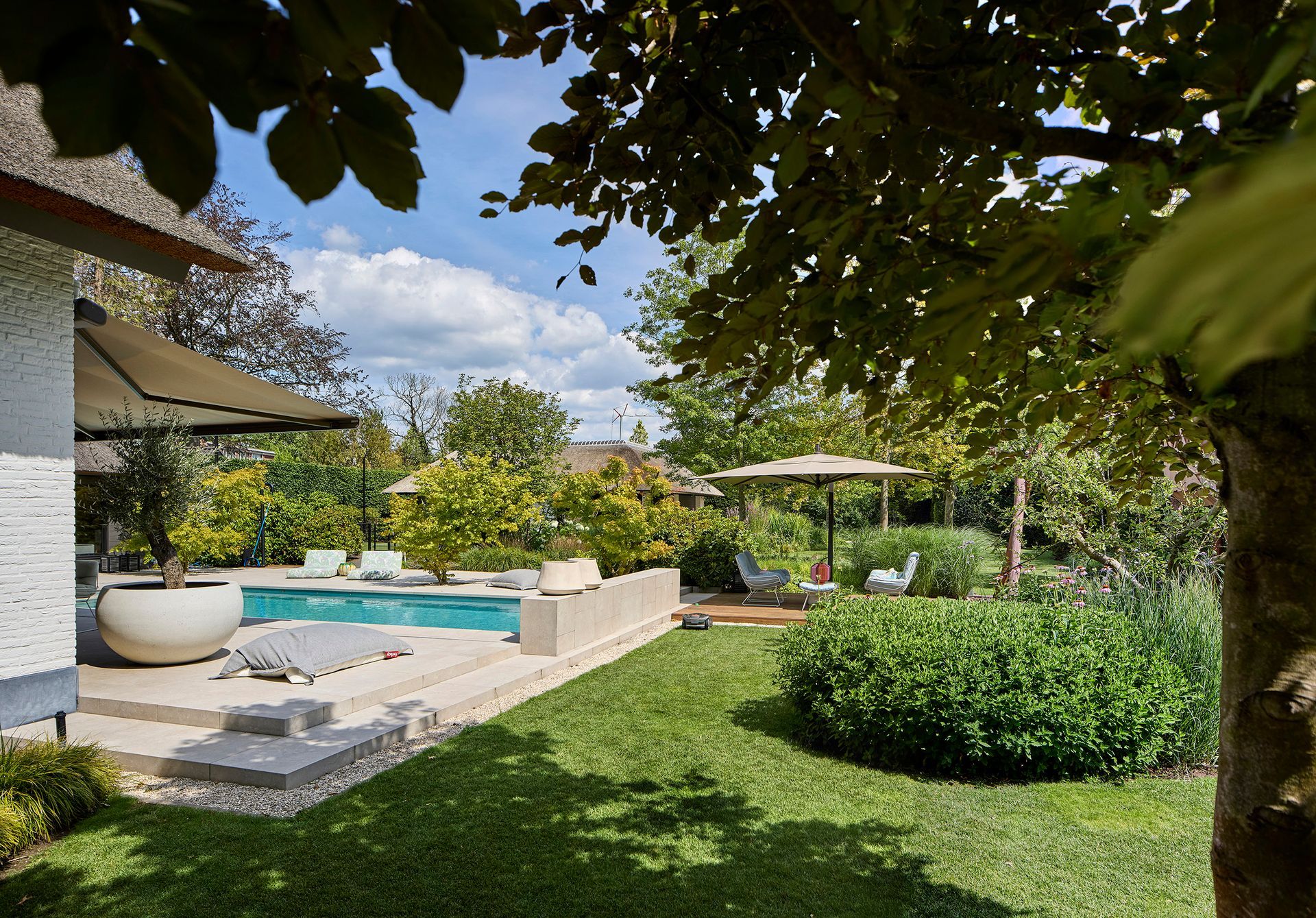 Moderne achtertuin met terras, turquoise zwembad, stenen loungehoek en weelderige groene tuin onder lommerrijke bomen.
