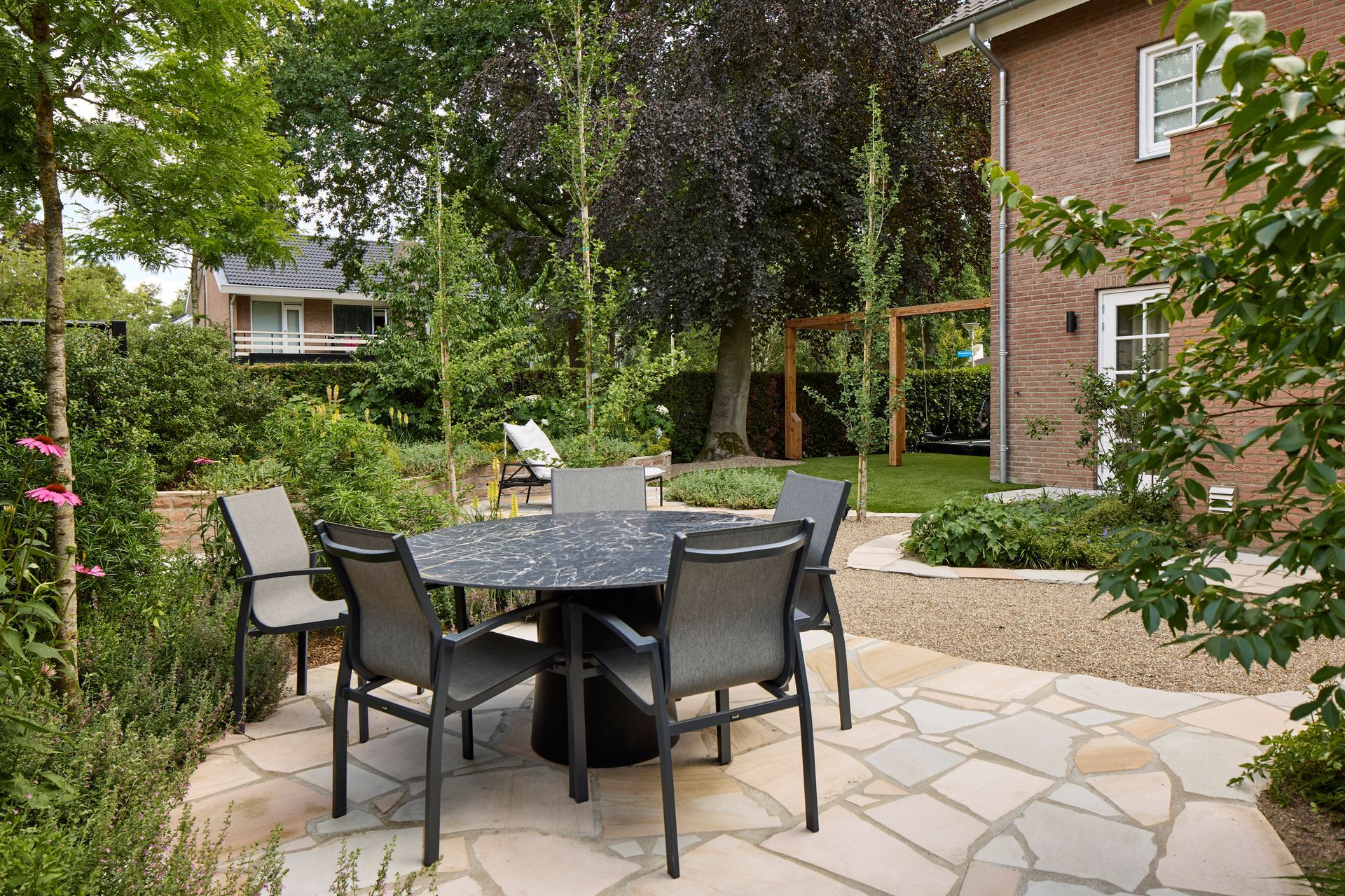 Terras met zwarte eettafel en stoelen op stenen bestrating, omgeven door groen naast een bakstenen huis.