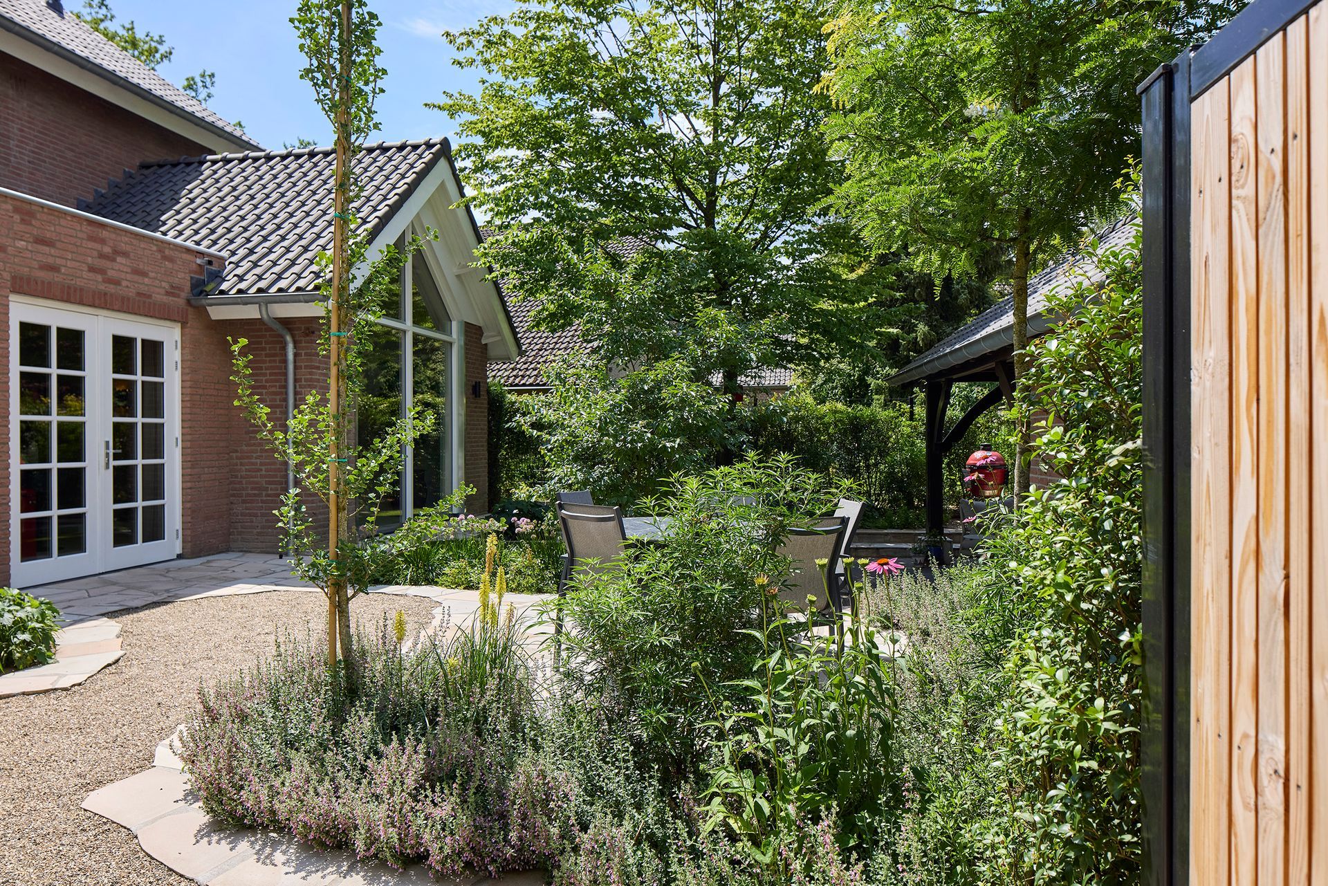 Een zonnige patio met bakstenen bestrating, struiken, bomen en een huis met openslaande deuren.