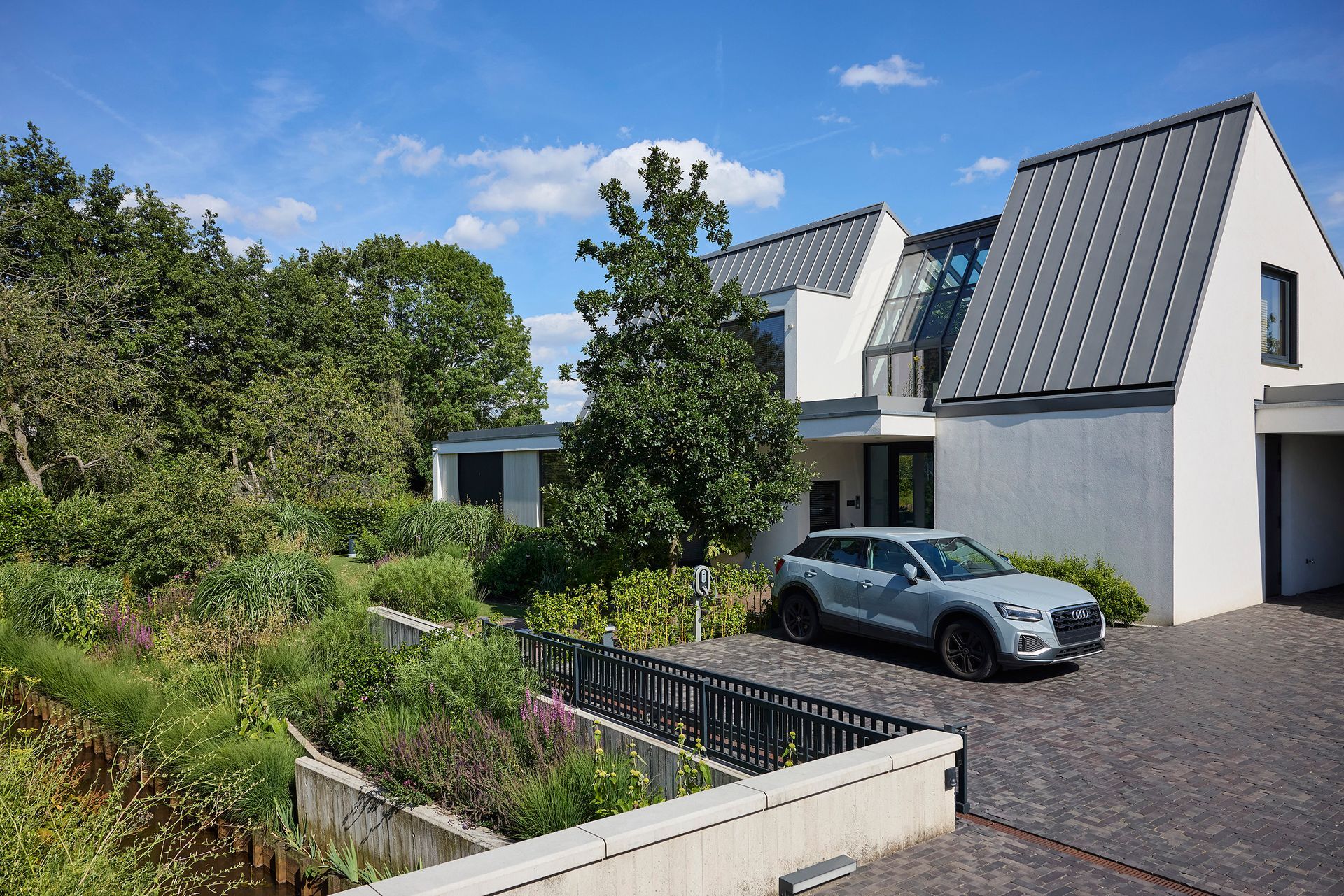Modern wit huis met donker dak, geparkeerde auto en weelderige tuin onder een blauwe hemel.