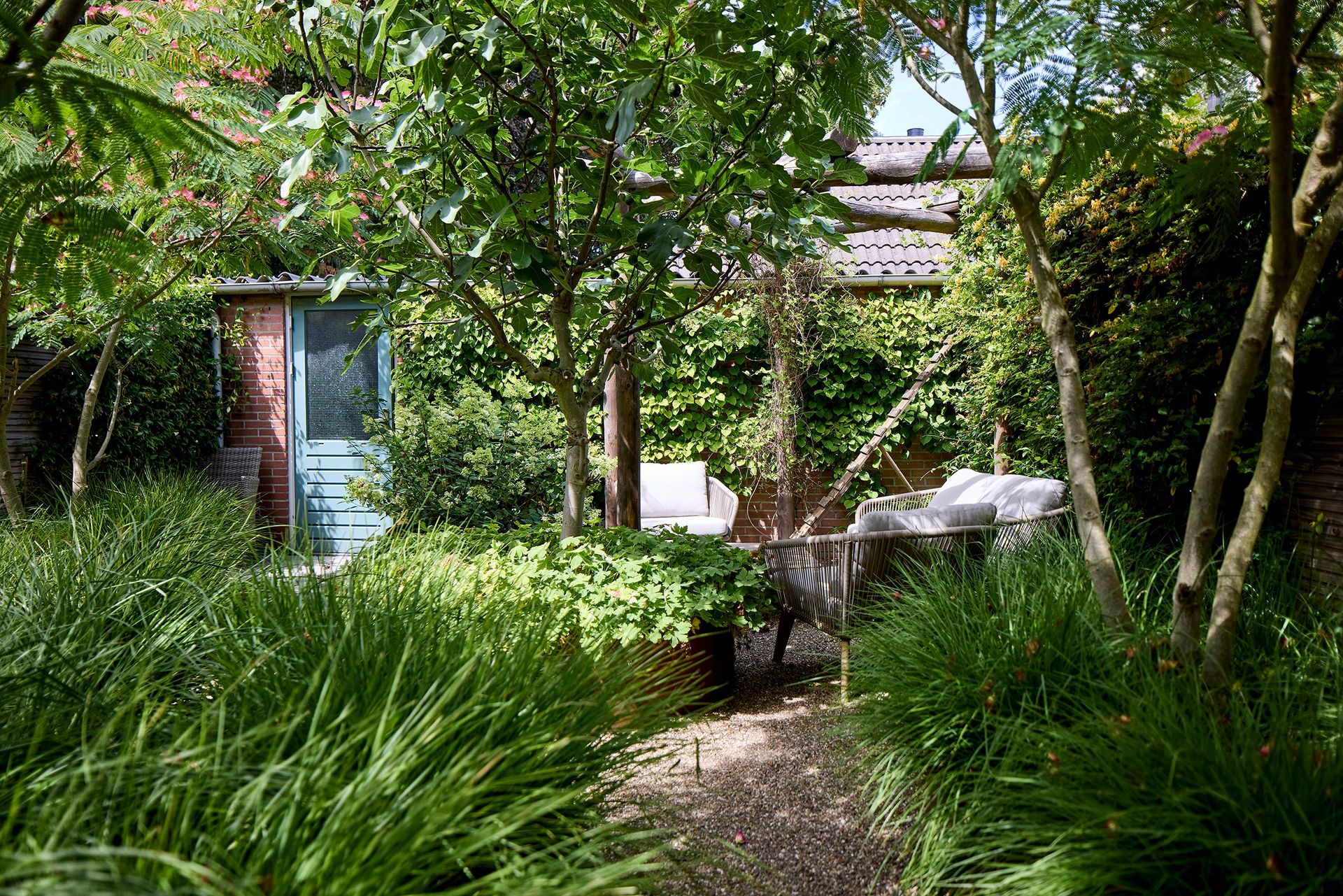 Een tuinpad naast een klein bakstenen huisje, omgeven door weelderig groen en bomen.